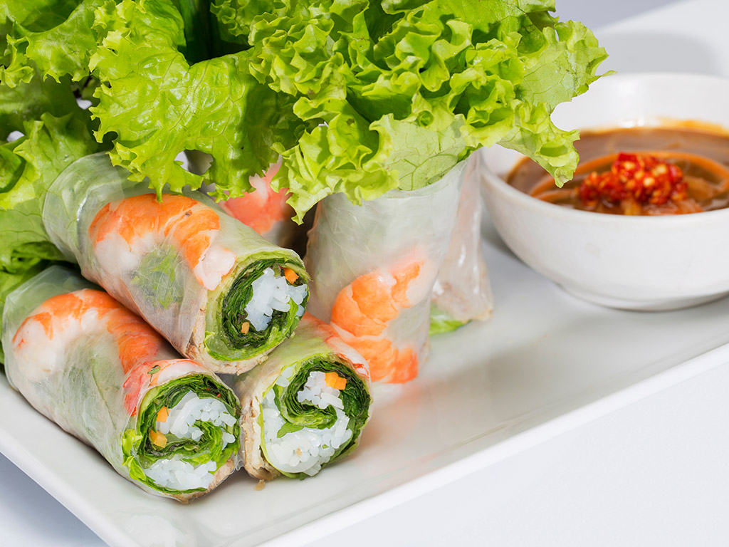 Goi Cuon is a dish that could come in both vegan and non-vegan versions. Each roll is wrapped inside rice papers and stuffed with green veggies such as lettuces, Vietnamese basils, rice noodles, and carrots. For meat-eaters, rolls can be added with sliced pork and shrimp.