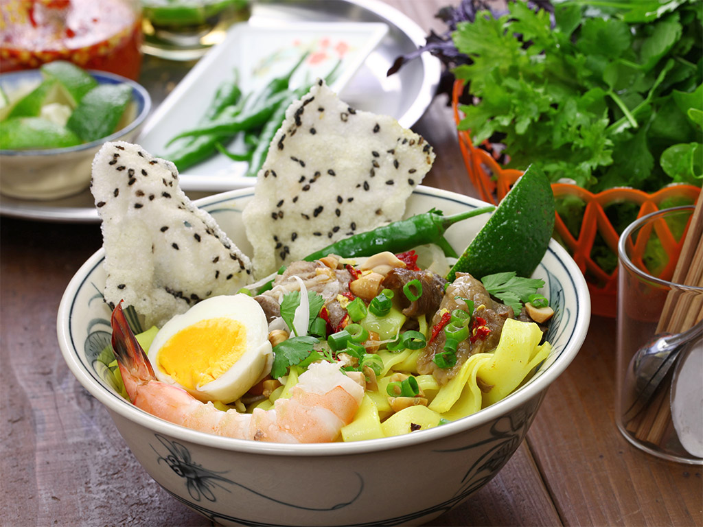 A traditional bowl of Mi Quang depicts the colour harmony of yellow noodles, white rice crackers, and green veggies.