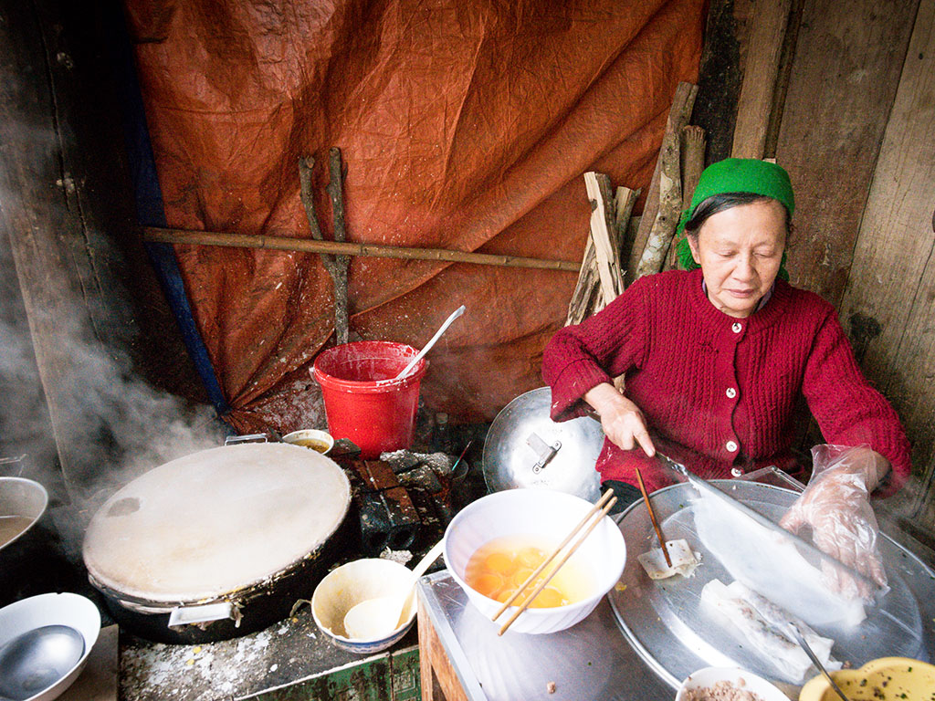 The cook uses a bamboo stick to remove the rice wrapper from the fabric and place it on a tray where it will be stuffed with the fillings.